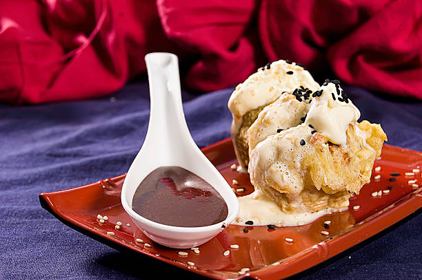 Close-up, tempura batter with vanilla ice cream on top, black and beige sesame seeds, ceramic spoon with liquid chocolate, rectangular wine-colored ceramic plate, on blue fabric, background, red fabric