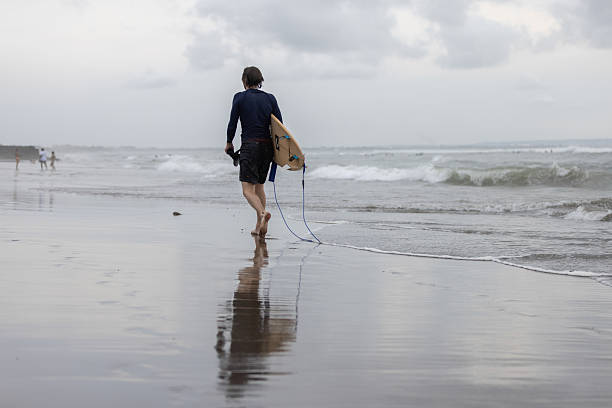 15 January 2025, Canggu Beach - Bali.A young surfer man walks along Canggu Beach in Bali carrying her surfboard, embodying the area’s vibrant surf culture and relaxed coastal spirit.