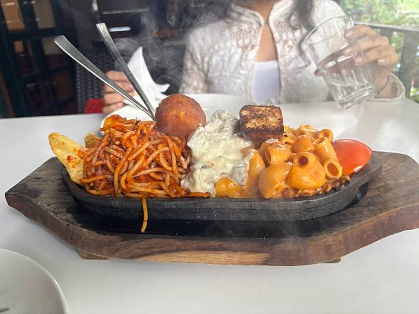 A close-up of a hot sizzler plate served in front of a hidden prince statue, featuring crispy cheese balls, spaghetti, and two varieties of pasta. Captured in warm lighting with rich textures and steam rising, ideal for food photography and culinary themes.