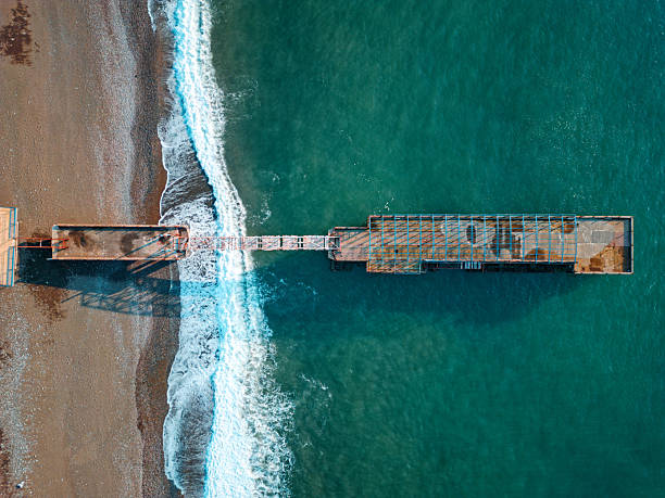 istock Top-down drone views of a weathered seaside pier: rusted metal frames stretch from a pebble beach into turquoise water as waves foam and shadows pattern the shore 2244368174