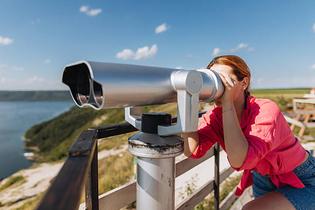 A woman in a pink shirt leans into a large silver telescope to gaze across a coastal landscape