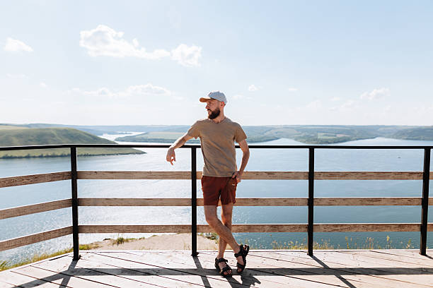 A relaxed man wearing a cap and casual clothes leans on a wooden railing, taking in expansive river views and rolling hills under a bright blue sky
