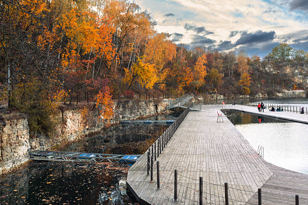 Krakow, Poland – October 23, 2025: Wooden walkways extend across a quiet quarry lake, surrounded by autumn trees and rocky cliffs. The mirrored surface shows soft evening light and cloud shapes.