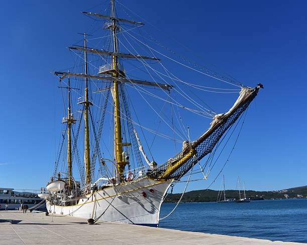A dramatic view of a grand, white-hulled tall ship docked prominently at the Budva harbor pier. The detailed rigging and multiple masts rise sharply against the cloudless blue sky, serving as a picturesque element of the port and highlighting maritime history and the tradition of large vessel sailing.