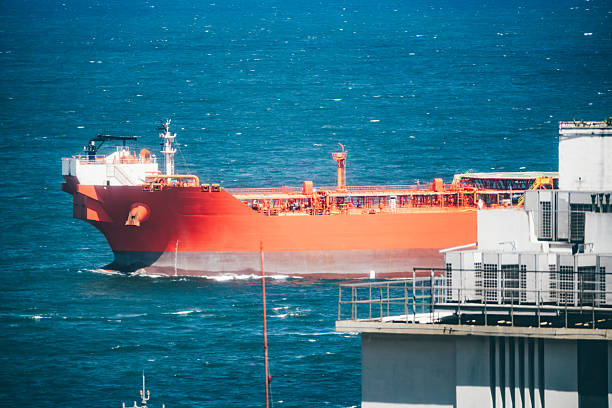 Large bright red oil tanker sailing in deep blue ocean water near an industrial port, captured with a telephoto lens showing ship details and modern building structures in foreground