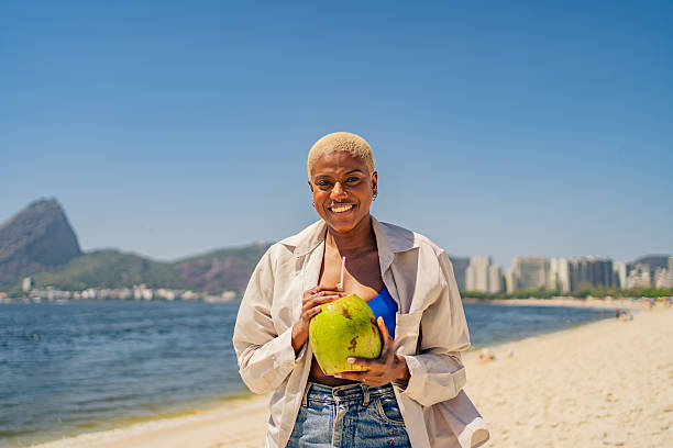 Smiling woman holding a fresh coconut drink on a sunny beach with mountain and cityscape in the background