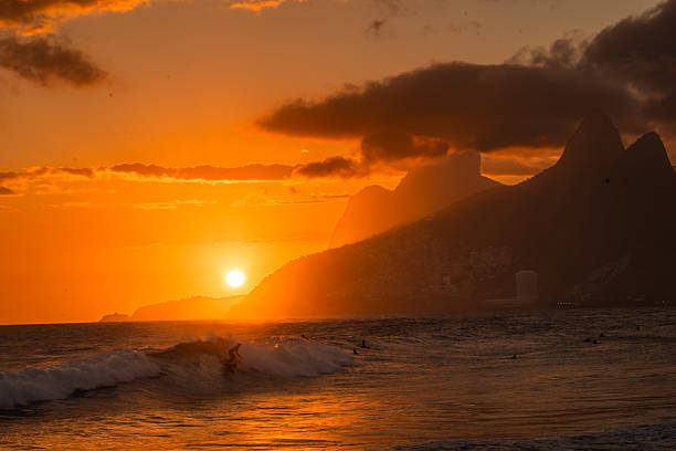 Surfer riding a wave at sunset with dramatic mountain silhouettes and orange sky over the ocean