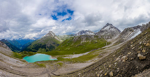 Scenic alpine landscape with a calm turquoise lake reflecting green grassy hills, dramatic rocky mountain peaks and drifting low clouds in bright summer weather, with wide blue sky and copy space.