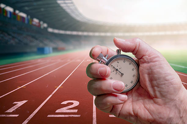 A close-up of a hand holding a stopwatch at a running track, symbolizing speed, timing, training, and athletic focus. The stadium and lanes provide context of competition and progress.