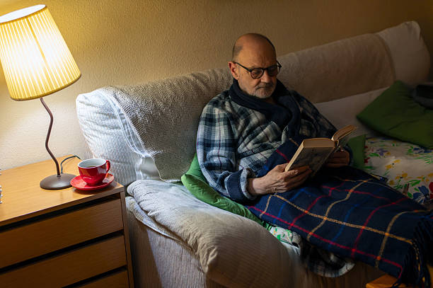 istock Older Man Reading Book on Sofa With Blanket, Lamp, and Cozy Home Vibe 2244418074