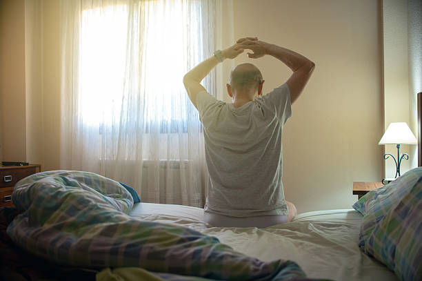 istock Man Sitting On Bed Stretching In Bedroom With Hands Raised At Morning Light 2244418100