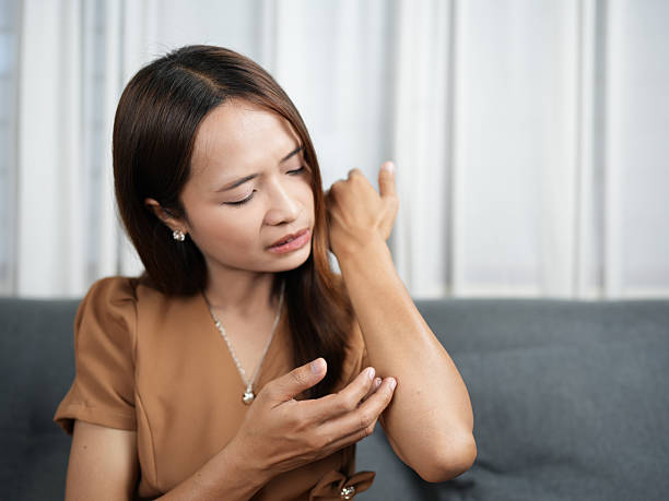 Woman brown blouse is sitting couch, looking concerned while examining her arm. She appears to be experiencing discomfort or irritation, showcasing moment of self care and attention to health