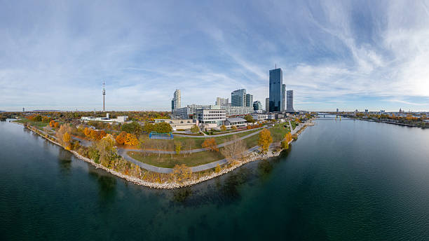 Aerial panoramic view of the Danube City (Donau City) skyline and the Danube Tower in Vienna, Austria, surrounded by colorful autumn foliage and the calm waters of the Danube River.