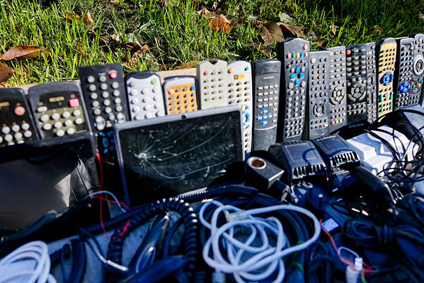 A large mixed lot of vintage TV and audio remote controls with tangled power adapters, old smartphones, batteries, and a worn tablet, showing heavy wear, scratches, dust, and missing buttons; many items appear retro, damaged, or non-functional, photographed outdoors on a market table.