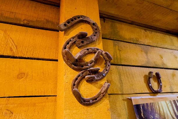 A set of old metal horseshoes displayed on a warm yellow wooden barn interior, including single and clustered pieces nailed or hanging on beams, with aged patina and cobwebs that emphasize rural craftsmanship and folk good‑luck symbolism.