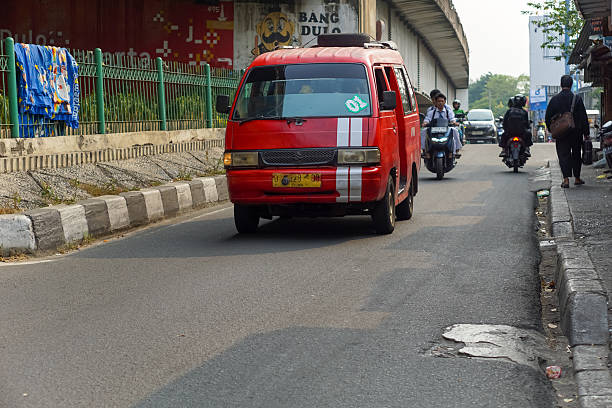 Jakarta, Indonesia - May 20, 2024: A red public minivan, locally known as “angkot,” drives along a city street under a flyover in Jakarta, Indonesia. The image captures daily urban traffic with motorcyclists and pedestrians.