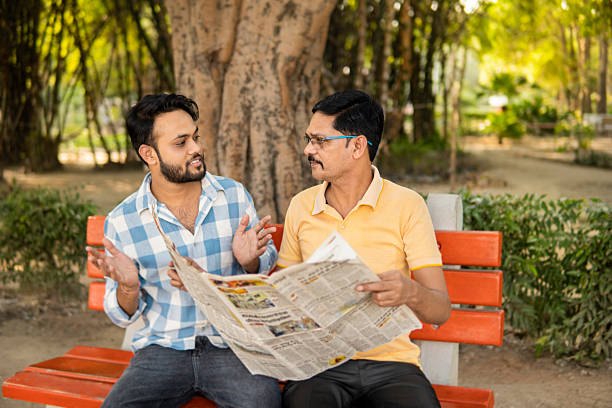 istock Mature Indian father and young son reading a newspaper together while sitting on park bench in fresh air. 2244432983