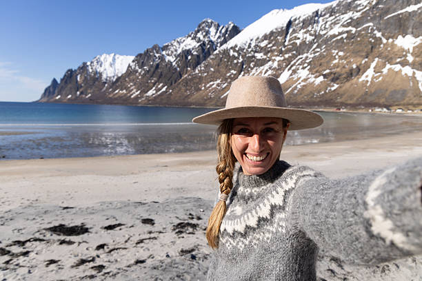 Smiling woman in wool sweater and hat taking selfie on snowy beach with mountains in background. Happy adult enjoying winter coastal landscape. Outdoor lifestyle and travel portrait.