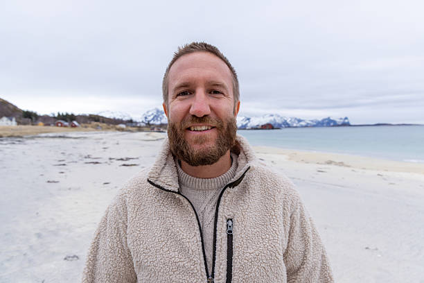 Bearded man in a cozy fleece jacket smiling on a cold beach with snowy mountains in the background. Happy outdoorsman enjoying winter coastal scenery. Portrait of a man in nature.
