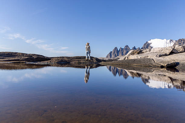 Adult woman in hat standing on rocky shore by calm water reflecting snowy mountains. Person enjoying solitude in nature with clear sky and rugged landscape. Outdoor adventure and peaceful moment.