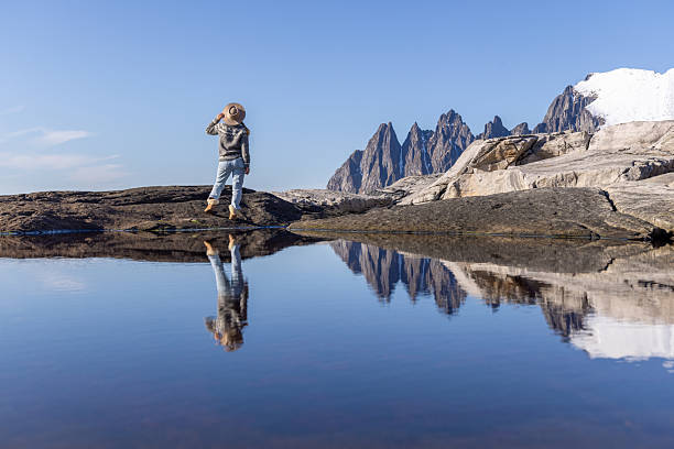 Woman wearing hat and sweater stands on rocky shore by calm water reflecting jagged mountains. Scenic outdoor landscape with clear sky and snow-capped peaks. Nature and solitude concept.