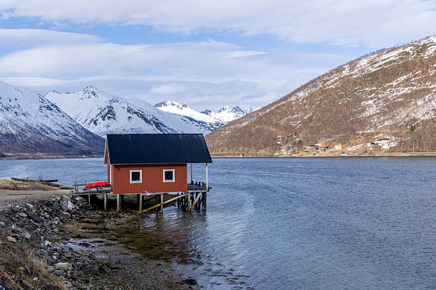 Small red cabin on wooden stilts by calm lake water with snowy mountains in the background. Scenic winter landscape with peaceful waterfront and rustic architecture. Mountain retreat by the lake.