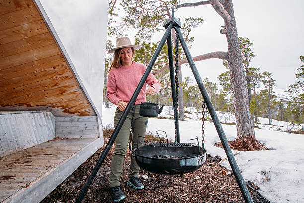 Woman in pink sweater and hat holding kettle near tripod grill outside snowy cabin. Outdoor cooking and camping scene in winter forest. Person setting up campfire cooking equipment.