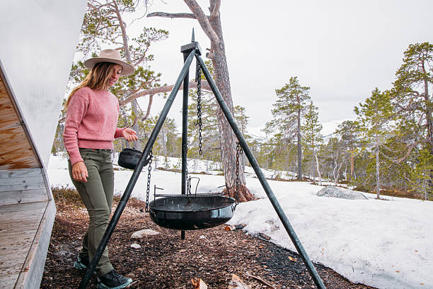Woman in pink sweater and hat holding kettle near tripod grill outside snowy cabin. Outdoor cooking and camping scene in winter forest. Person setting up campfire cooking equipment.