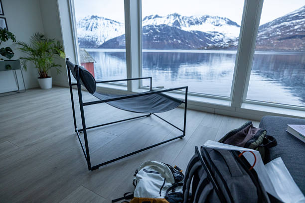 Minimalist black metal chair near large window overlooking snowy mountains and calm fjord. Cozy interior with backpacks and plants, perfect for relaxation or remote work. Scenic nature view from indoors.