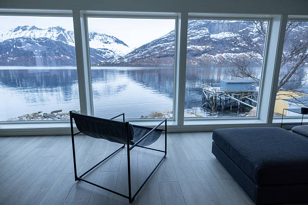 Minimalist living room with black chair and ottoman facing large windows. Scenic fjord and snow-covered mountains visible outside. Cozy interior with natural light and nature backdrop.
