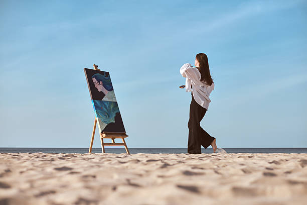 Young female artist paints by sea, capturing inspiration from coastal setting. Scene reflects tranquility and creativity with sandy beaches and blue skies enhancing artistic ambiance