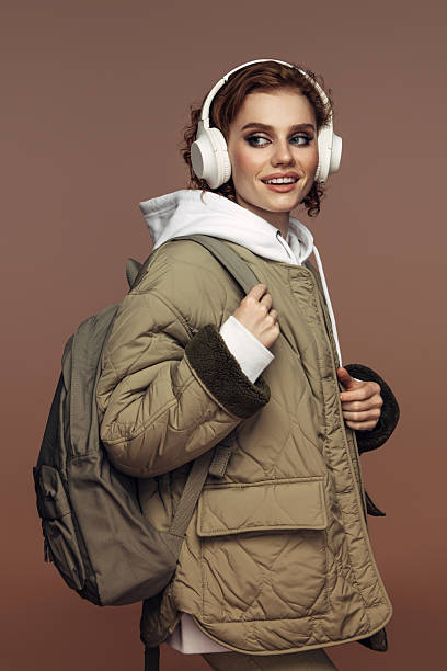 A young woman in a casual, urban outfit is listening to music through large, over-ear headphones. She is dressed in a cozy, oversized jacket with fur-trimmed cuffs and a white hoodie underneath. Carrying a large backpack, she looks over her shoulder with a cheerful smile, exuding a relaxed and trendy vibe. The warm, neutral background complements her laid-back yet stylish appearance, making this image perfect for conveying themes of modern lifestyle, music, and travel.Keywords: