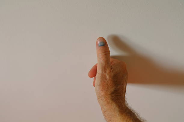 A male hand with a blue thumbnail is pointing towards or pressing against a white wall. The hand is positioned centrally with ambient lighting casting a soft shadow. The wall appears smooth, and the hand shows signs of maturity with visible veins and skin texture.