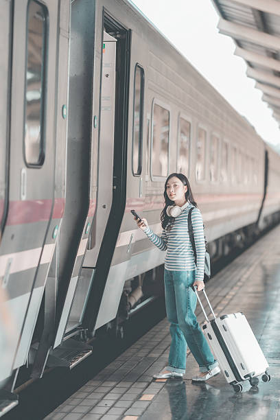 istock Asian woman with backpack looking forward while standing at train station, holding luggage, waiting for train to depart at busy station, excited about the journey. 2244475756