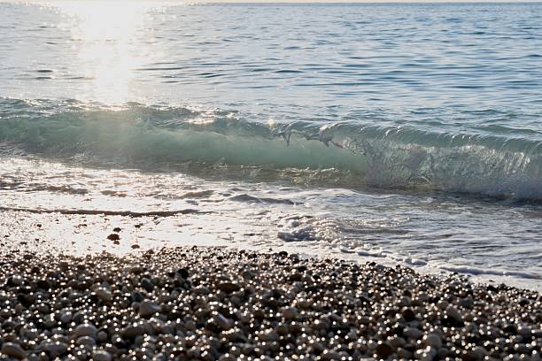 A dynamic close-up of a breaking sea wave creating white foam over smooth, multi-colored pebbles on a beach, highlighting the natural texture and motion.
