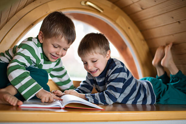 Two young boys wearing striped long-sleeve shirts lie on their stomachs on a wide window sill, sharing a book together. Smiling as they look down at the open book between them, with one boy pointing at the pages. Behind them, a large circular window with wooden framing lets in bright natural light.Shot with Nikon D800