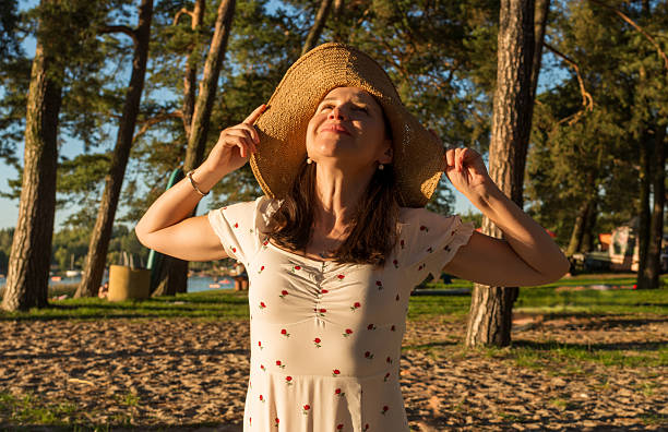 Joyful woman lifts her face toward the sun, smiling warmly under a wide straw hat. Surrounded by tall trees and soft golden light, she captures the carefree spirit of a summer afternoon.