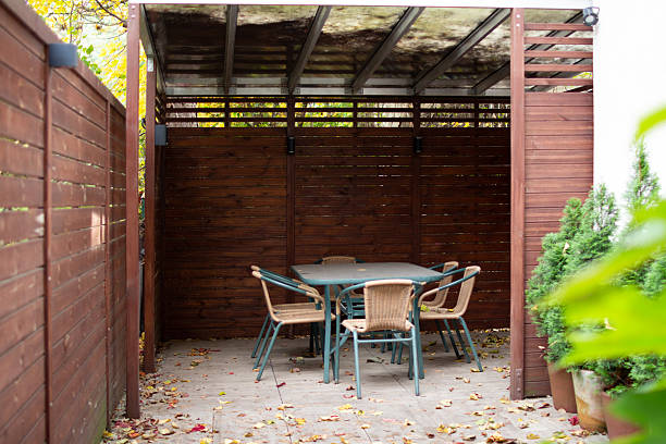 Unoccupied exterior space with ivy at stepped entrance, pale gray L-shaped sofa and pillows, portable fire pit, and slatted ceiling providing partial shade.