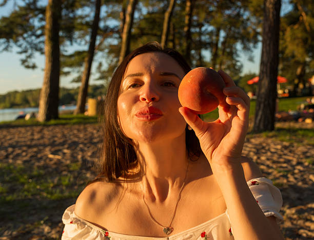 Cheerful woman in a floral dress holds a ripe peach close to her face, glowing in warm evening light. The soft background of trees and sand enhances the joyful, relaxed summer feeling.