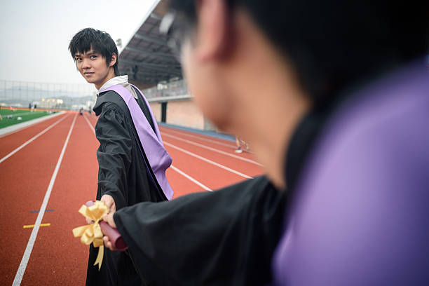 Two young Asian men in graduation gowns and stoles, standing on a running track at an athletic stadium is a precise and accurate depiction. Symbolizing the passing of the torch or the completion of a long race/journey\