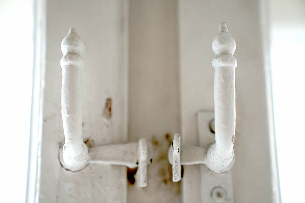 Macro shot of a symmetrical vintage latch, painted white, with decorative curved posts. A detail of old hardware on a light-colored wall or frame, emphasizing the antique decor and the need for renovation.