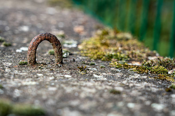 Low-angle close-up of a rustic, rusty metal loop embedded in gray concrete or stone, with patches of moss and a brightly blurred green background on the right
