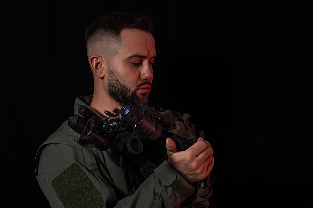 Serious man in tactical gear holding a helmet with night vision goggles, featuring two monoculars connected by a bridge.