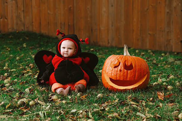 An adorable baby girl, eleven months old, sitting in the grass with a jack o'lantern. She is dressed in a ladybug costume for halloween. Shot with a Canon 5D Mark IV.