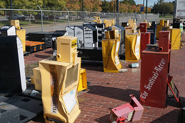A collection of discarded newspaper vending machines stands on brick pavement near a chain-link fence in Little Rock, Arkansas. Labeled with titles like Democrat Gazette and The Sentinel-Record, the faded and damaged units evoke themes of media obsolescence, regional press legacy, and the decline of physical distribution. Little Rock, AR, USA on October 29, 2025