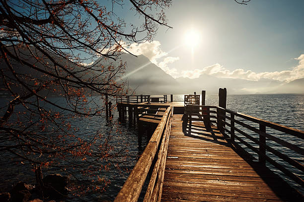 istock Scenic wooden pier extending over tranquil water, surrounded by mountains and trees, illuminated by warm sunlight, creating a serene atmosphere 2244525200