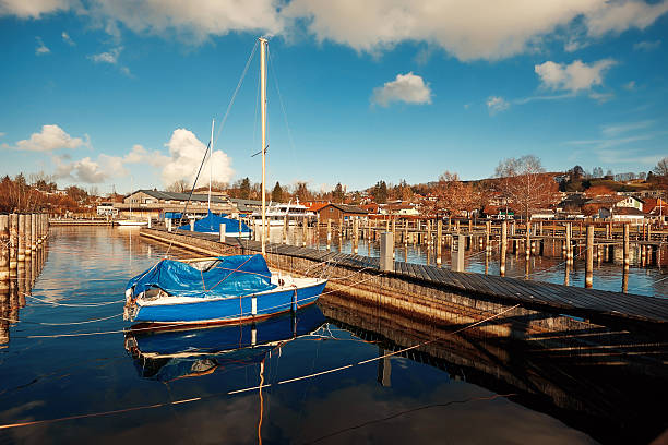 Blue sailboat is anchored in a tranquil marina, with reflections on still water and a picturesque sky filled with fluffy clouds, creating a peaceful atmosphere