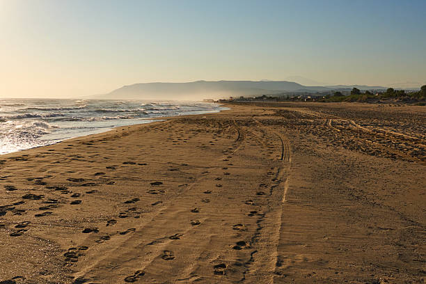 istock Tranquil beach scene at sunset with gentle waves lapping against sandy shore, showcasing footprints and distant mountains in the background 2244528212