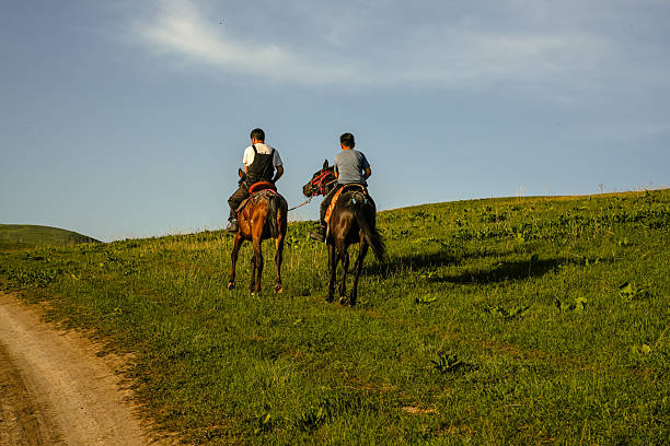 Two men ride horseback uphill along a narrow dirt trail surrounded by lush green grass in the mountain pastures of Central Asia. The scene captures a typical rural landscape of Kyrgyzstan, where horseback travel remains a traditional and practical means of moving across wide, open highland terrain. Evening sunlight casts long shadows and warm tones over the hills, emphasizing the vastness and peaceful atmosphere of the countryside. This image highlights authentic daily life in the region, showcasing local riders, traditional saddles, and the pastoral environment characteristic of Central Asian nomadic culture.Kyrgyzstan, Besh-Kungey, - May 03 2025