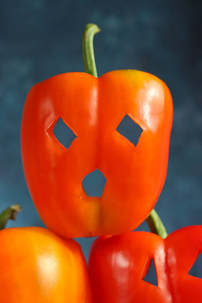 Stock photo showing close-up view of three, orange and red bell peppers carved like Jack O'Lanterns stacked in a pyramid against a mottled blue-green background.